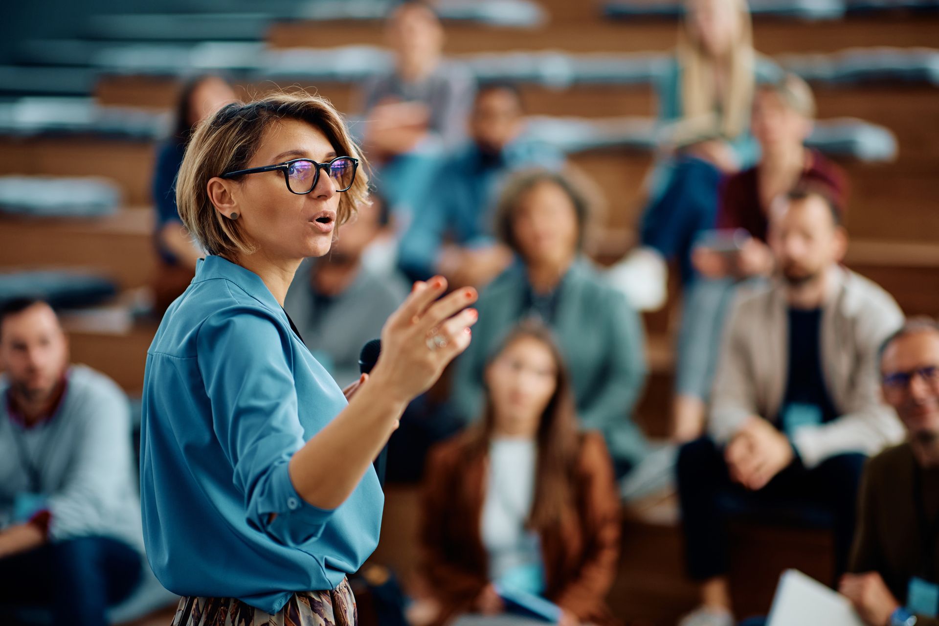 A speaker in a blue shirt gestures while presenting to an audience seated in a tiered auditorium.