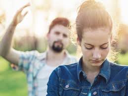 A person with an upset expression looking down, while someone behind them raises a hand in the background outdoors.