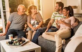 Four people sit on a couch in a bright living room, laughing and conversing while a bowl of snacks sits on a table.