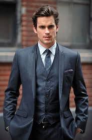 A man wearing a dark three-piece suit, light shirt, and tie, standing outdoors in front of a brick building.