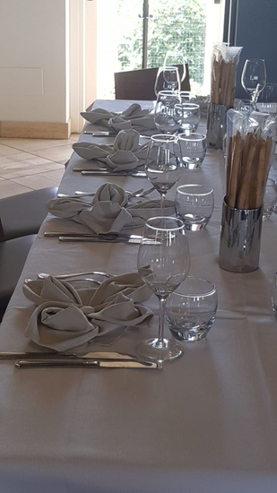 Un lungo tavolo apparecchiato per un pasto con tovaglioli piegati, bicchieri, posate e un barattolo di grissini accanto a una porta luminosa