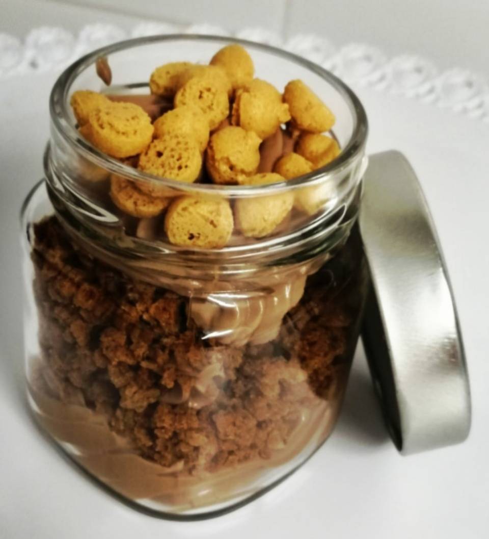 Barattolo di vetro aperto contenente granola con pezzetti gialli in cima, coperchio appoggiato di fianco su una superficie bianca.