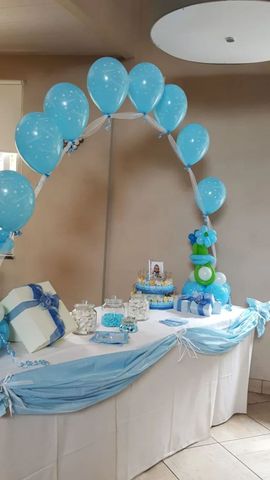 Arco di palloncini blu sopra un tavolo decorato con bomboniere blu e bianche e torta, sullo sfondo di una parete semplice.