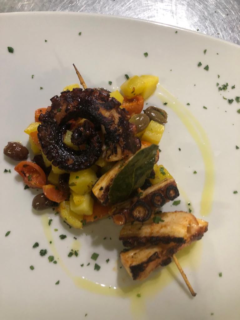Piatto gourmet impiattato con carne grigliata, verdure e guarnizione di erbe aromatiche su un piatto bianco.