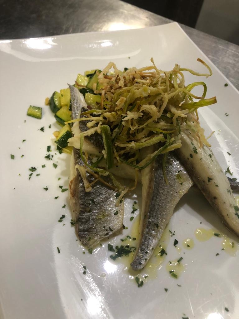 Piccoli pesci argentati serviti su un piatto bianco, guarniti con verdure fritte croccanti ed erbe aromatiche.