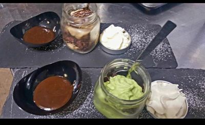 Tavolo dei dolci con salsa al cioccolato, vasetti di frutta, panna montata e mousse verde su un piano di lavoro scuro.