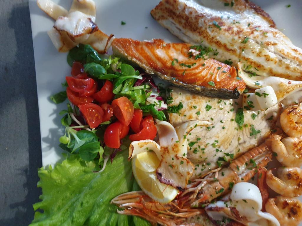 Salmone grigliato con pomodorini, verdure, spicchi di limone, gamberi ed erbe aromatiche su un piatto bianco.