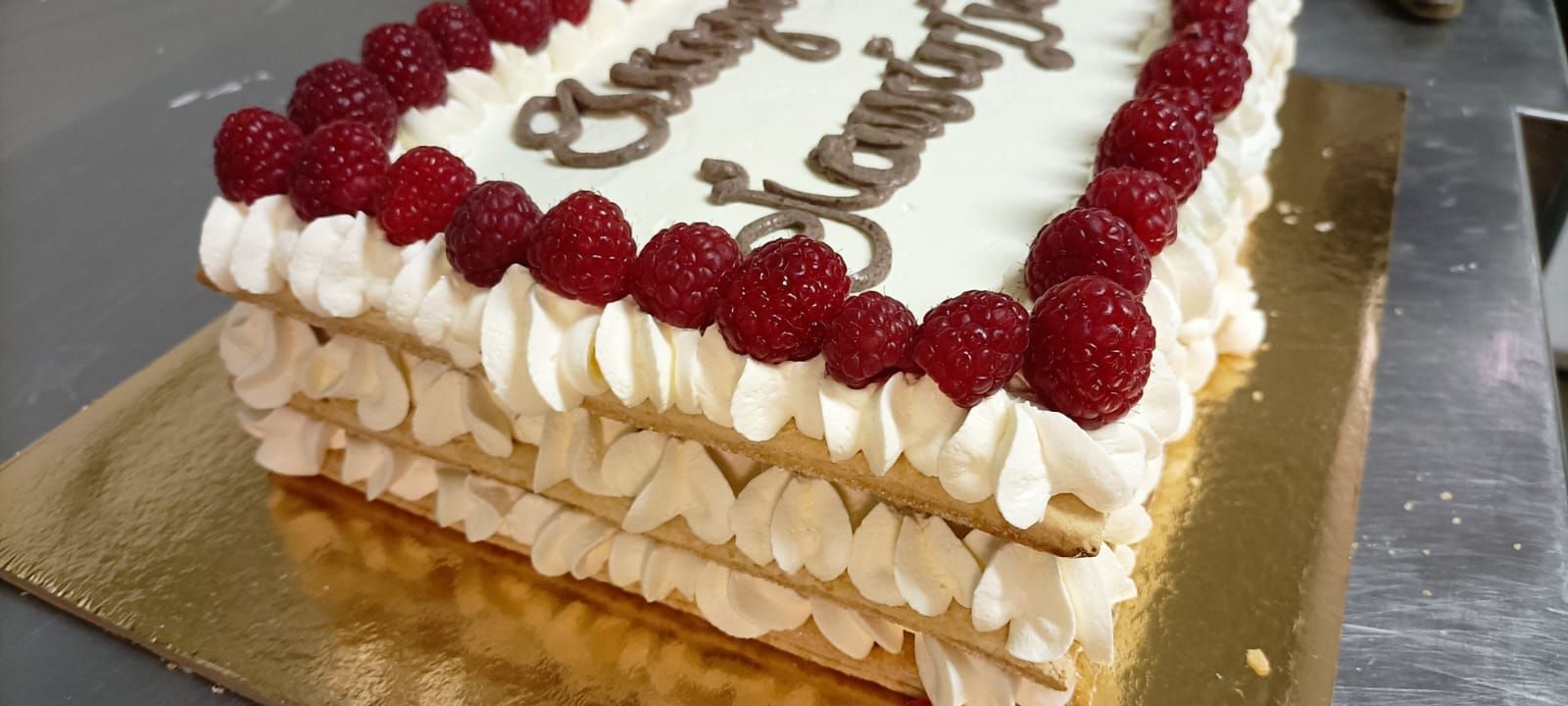 Torta rettangolare con glassa bianca, lamponi e scritta in cioccolato su una base dorata.