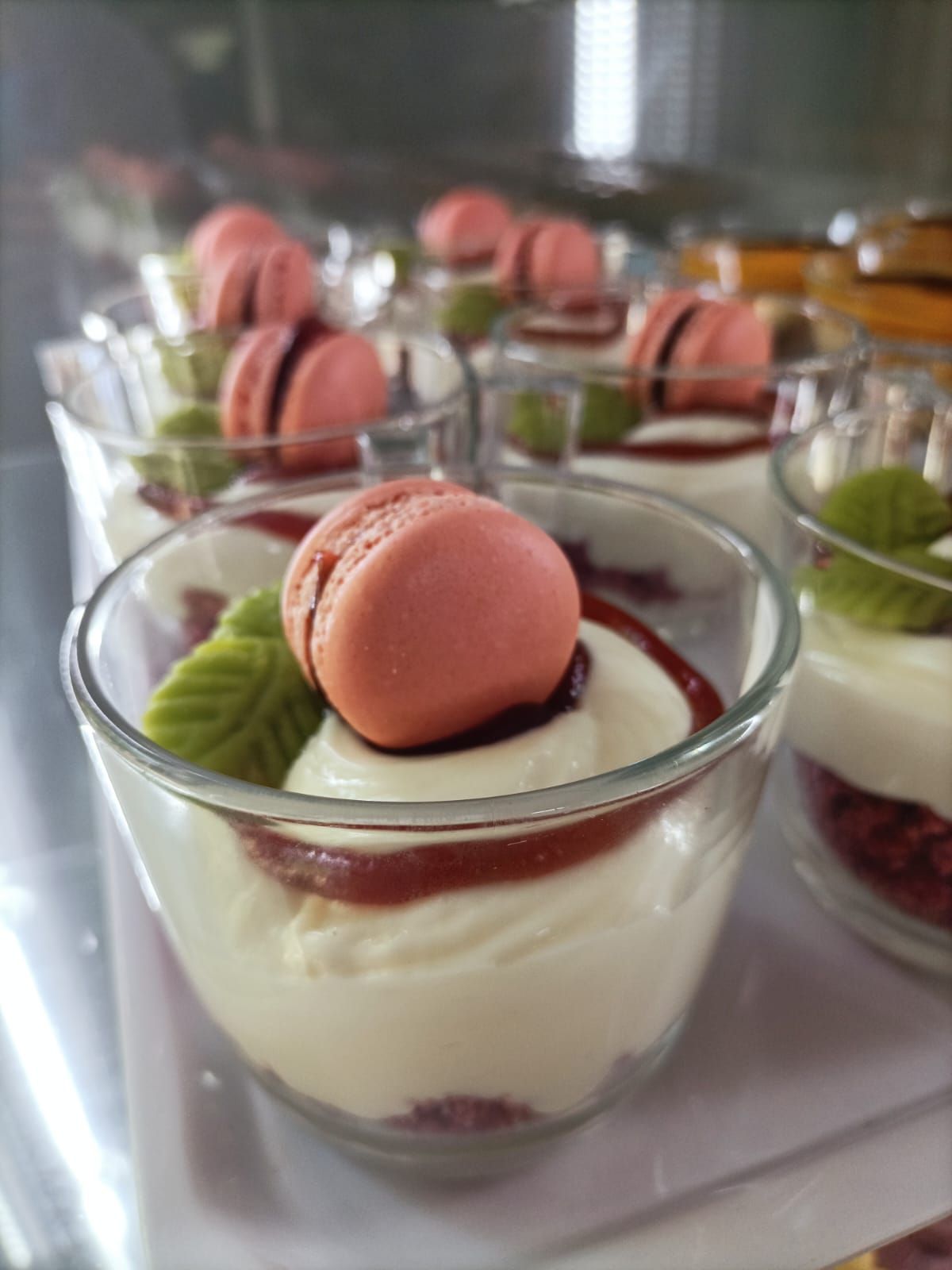 Bicchieri di dessert a strati guarniti con macarons rosa e verdi su un vassoio bianco.