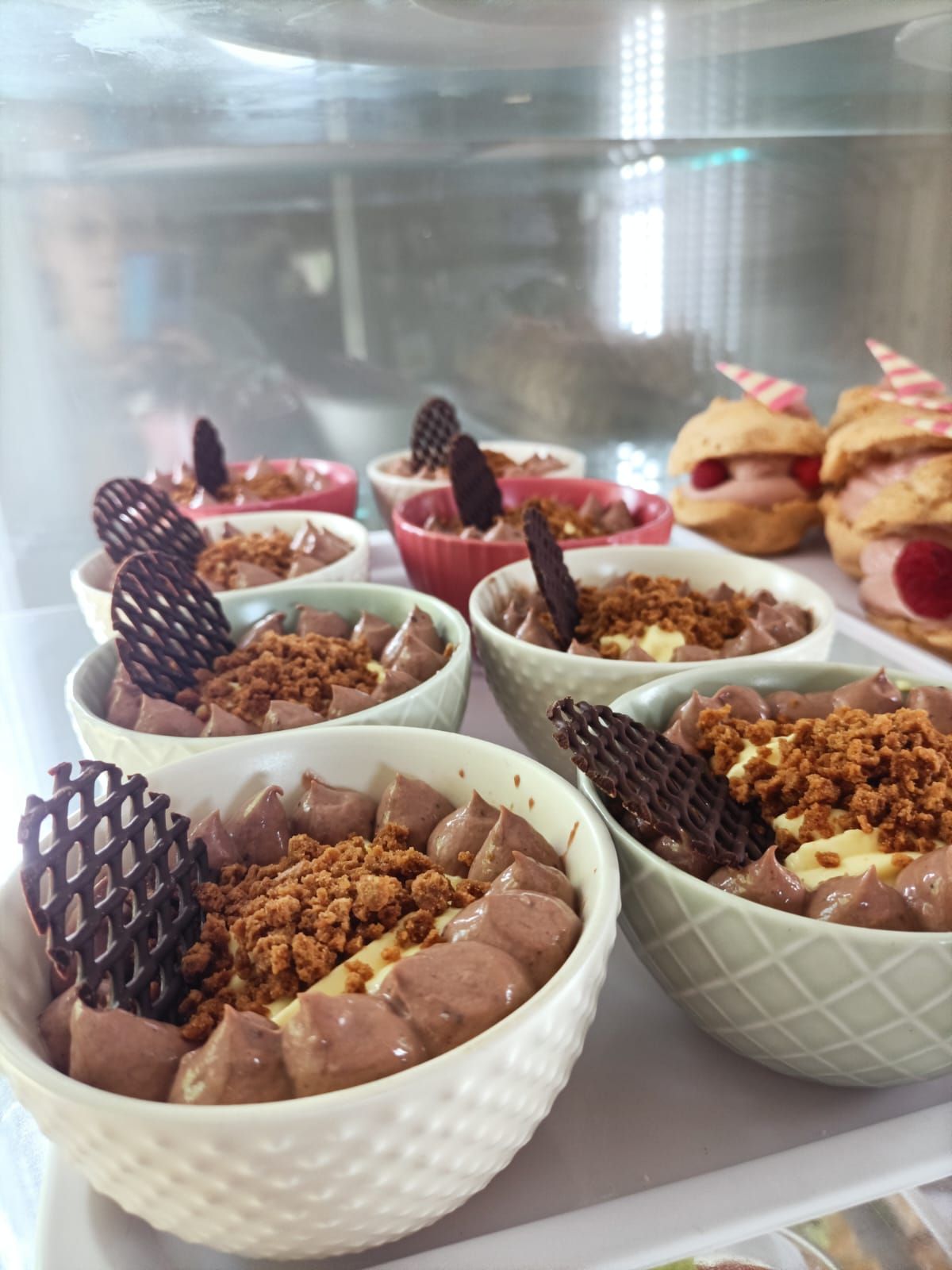 Un assortimento di ciotole da dessert con guarnizioni di cioccolato e granola su un vassoio espositivo, con pasticcini sullo sfondo.