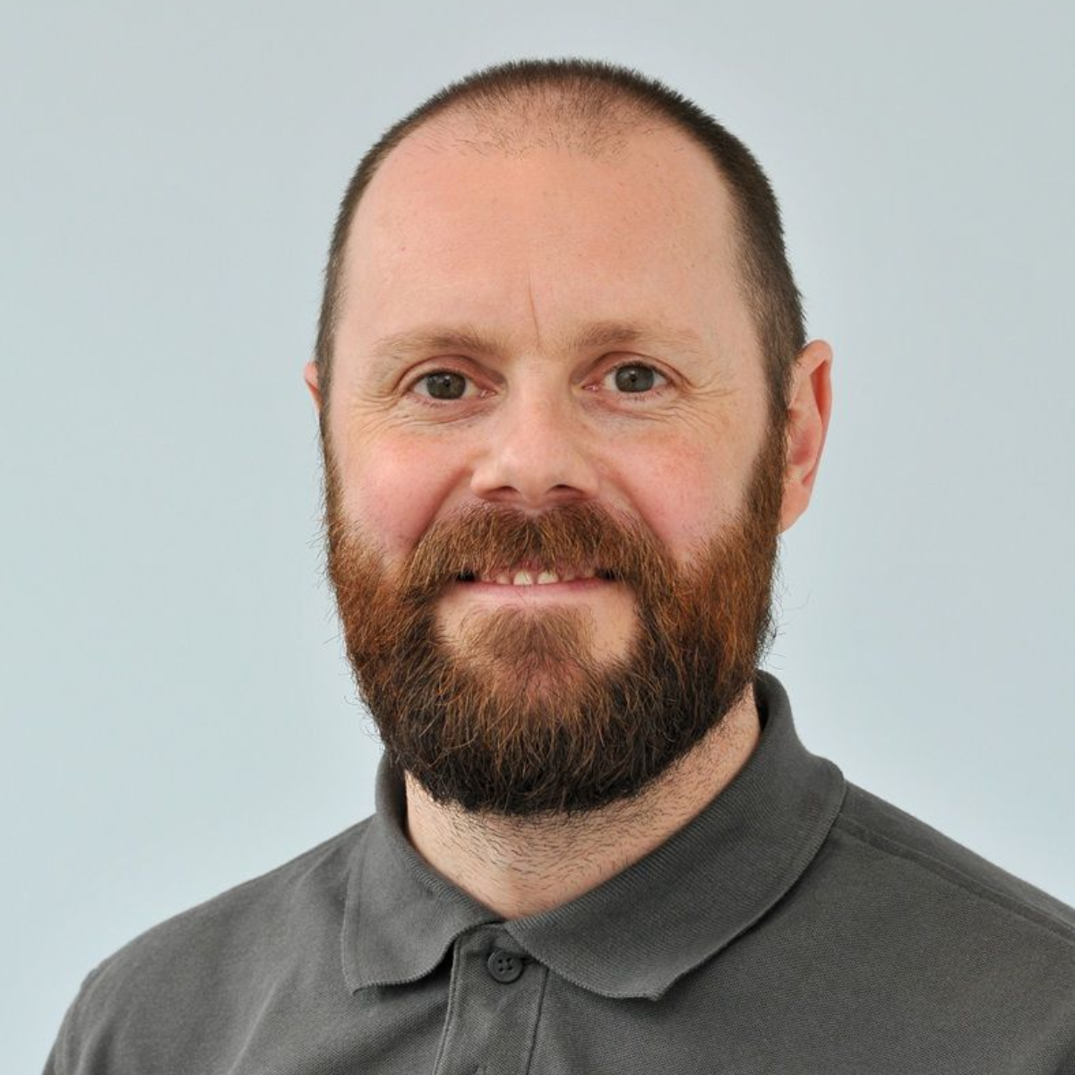 Portrait of a smiling man with a beard wearing a gray collared shirt against a light background
