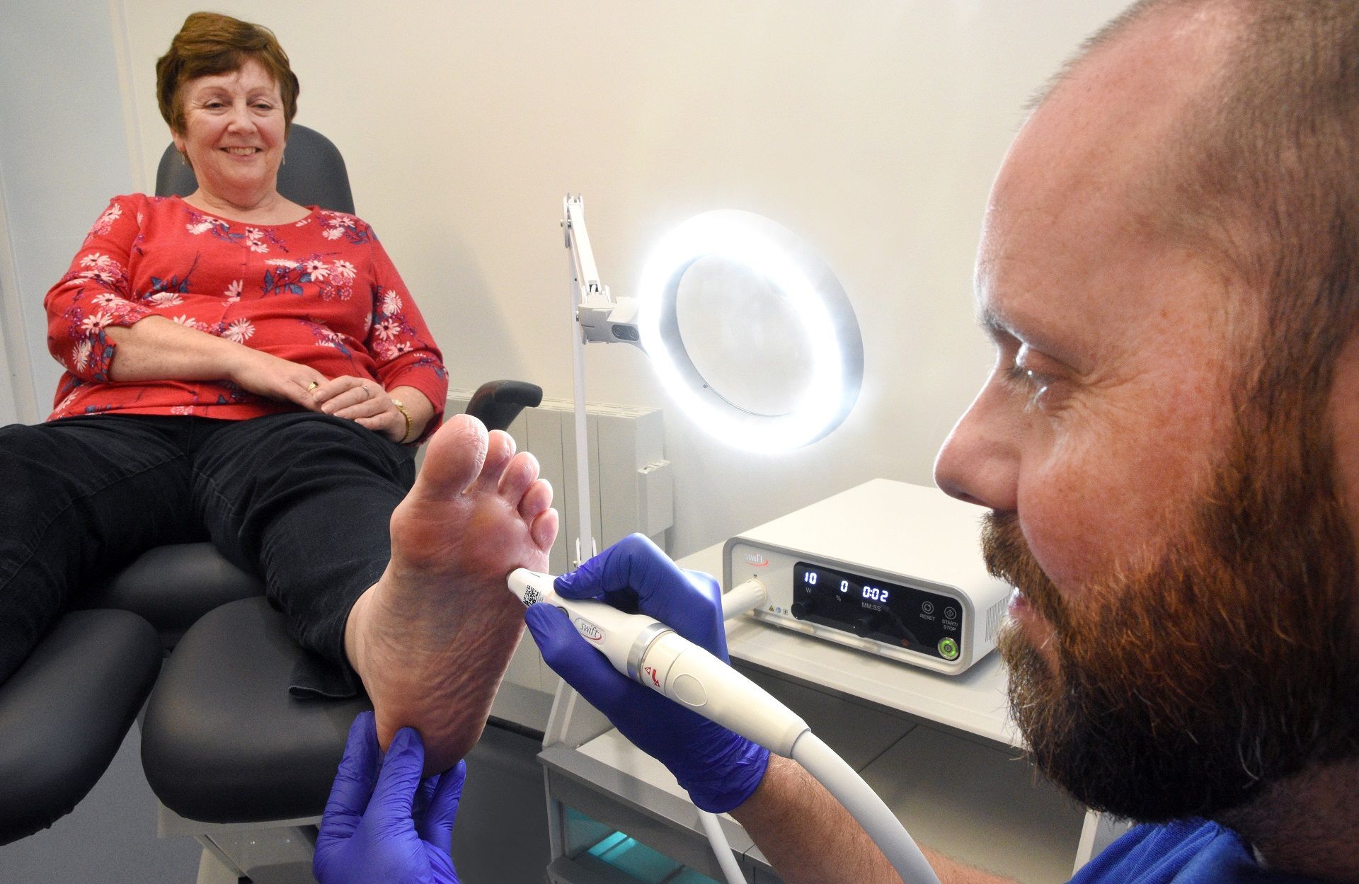 Foot care treatment in a clinic, with a clinician examining a patient’s toes under a ring light.