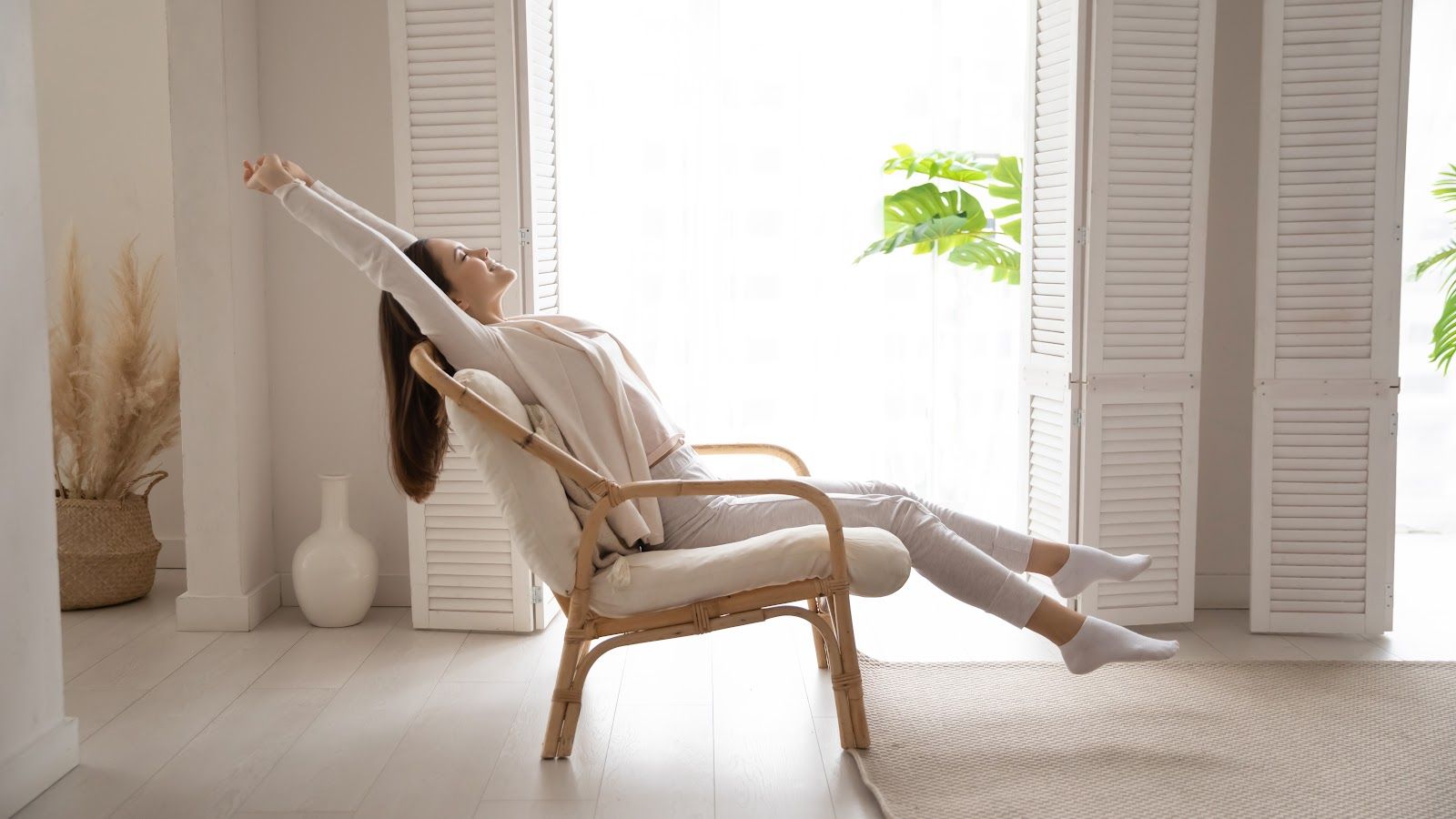 Person reclining on a wicker chair in a bright, minimalist room, stretching with arms extended.