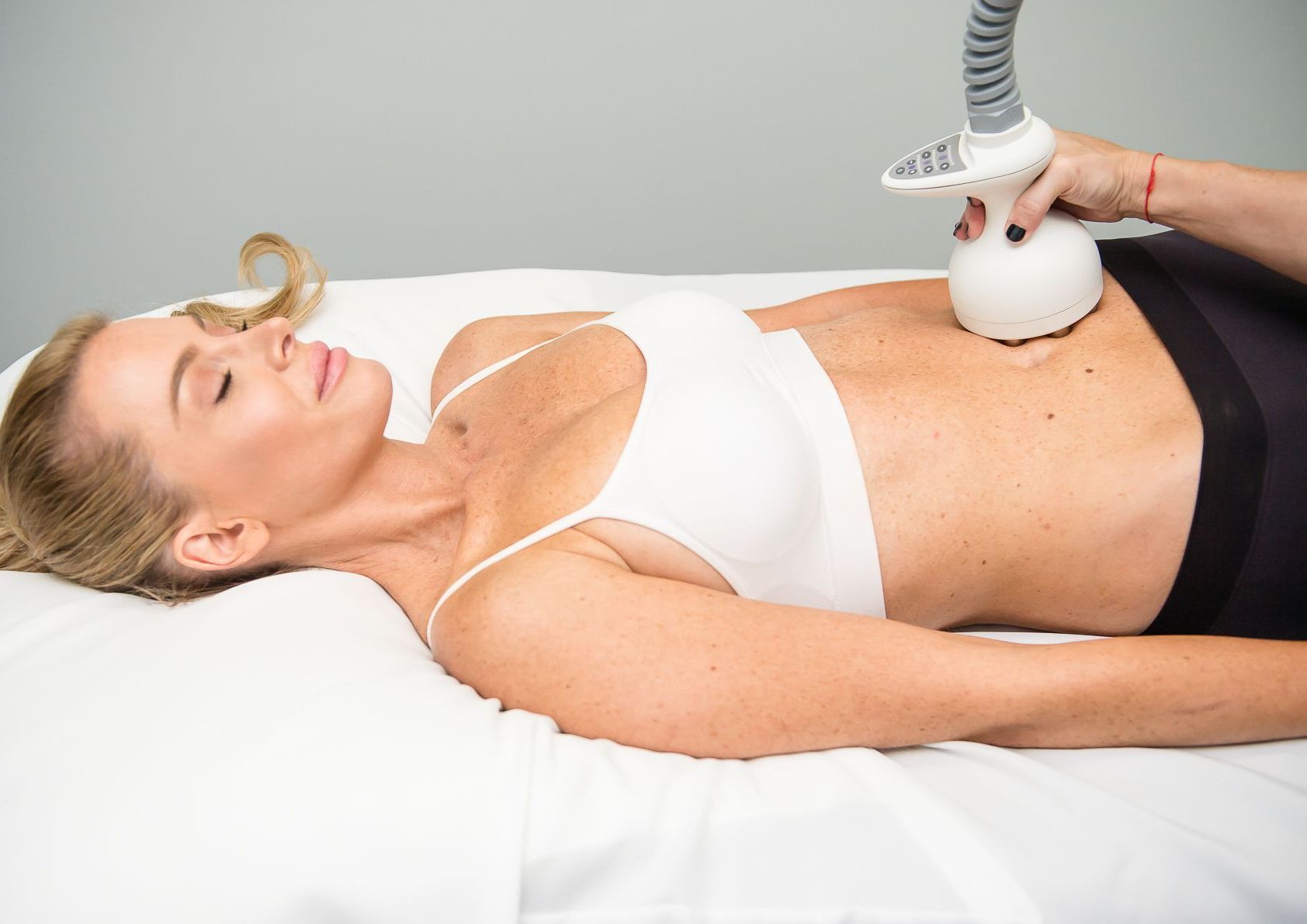 A woman is laying on a bed getting a cavitation treatment on her stomach.