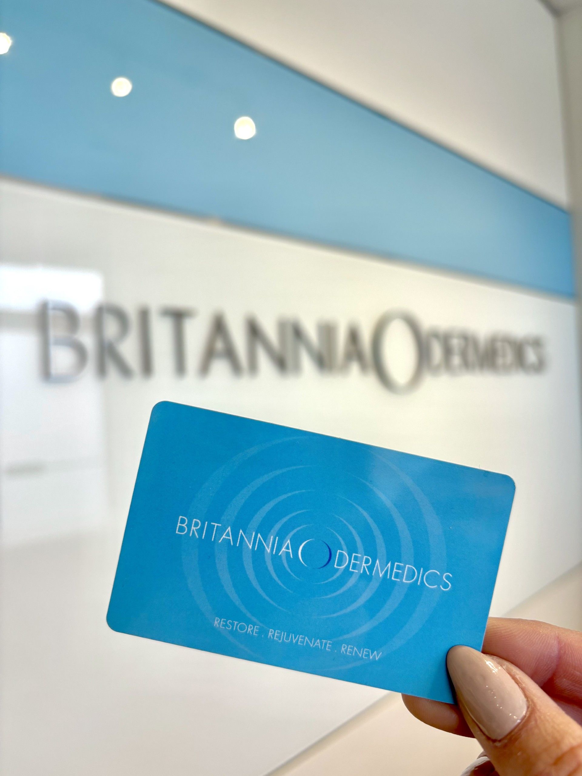 Blue card held up in front of a blurred Britannia Dermis sign in a clinic lobby