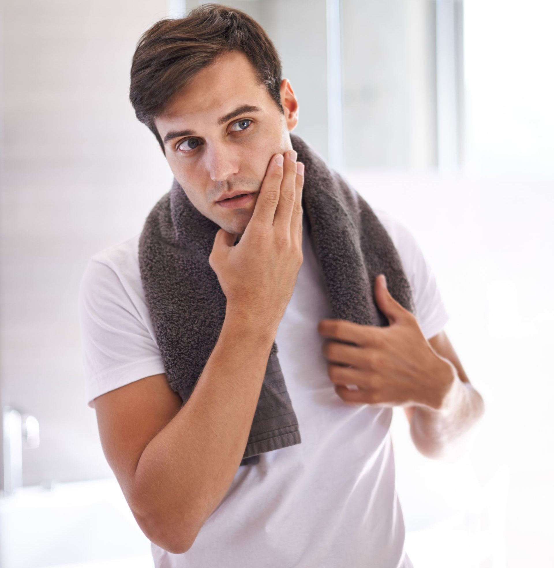 A man is looking at his face in the mirror while holding a towel around his neck.