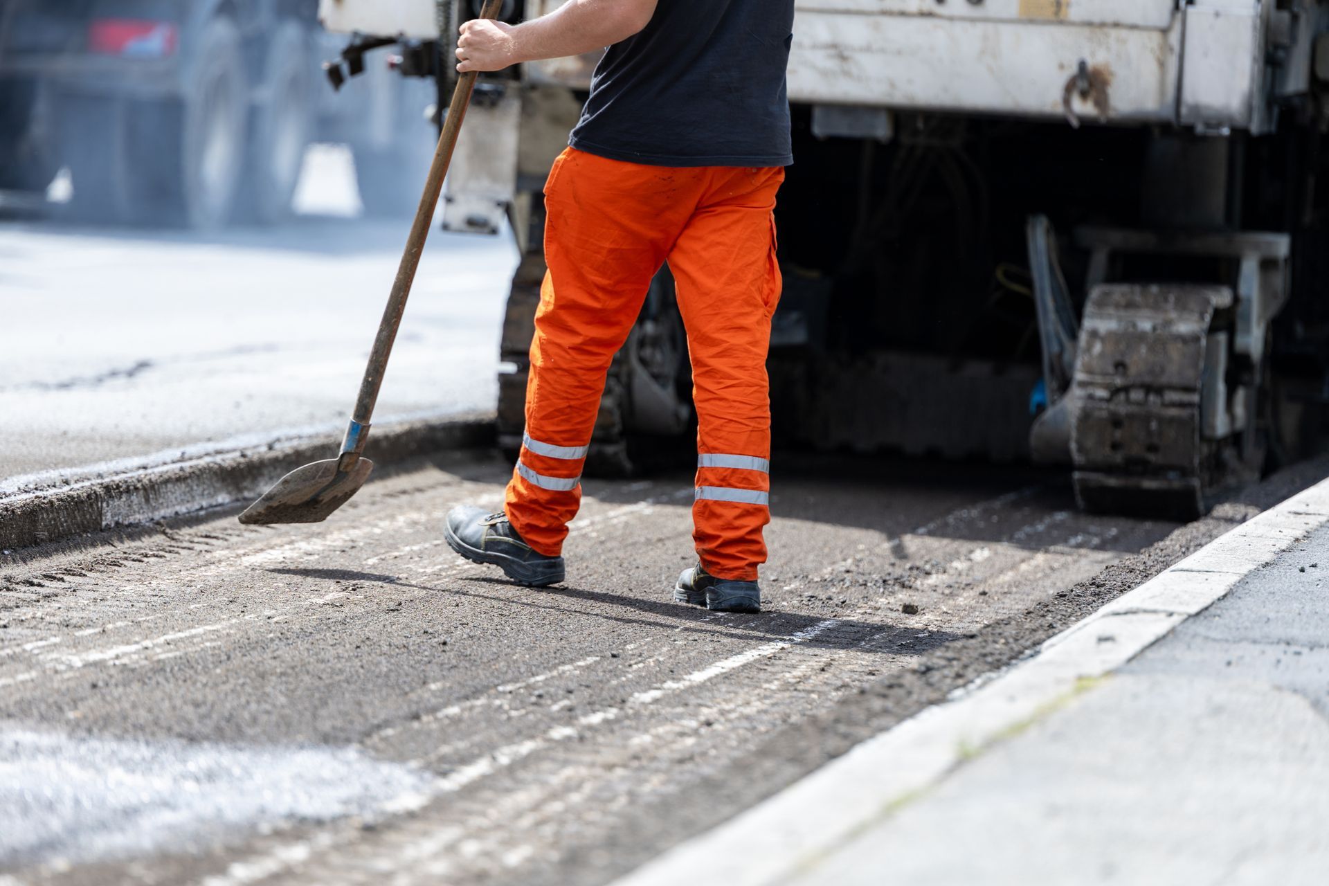 Worker using a shovel during asphalt repaving next to heavy machinery on a city street.