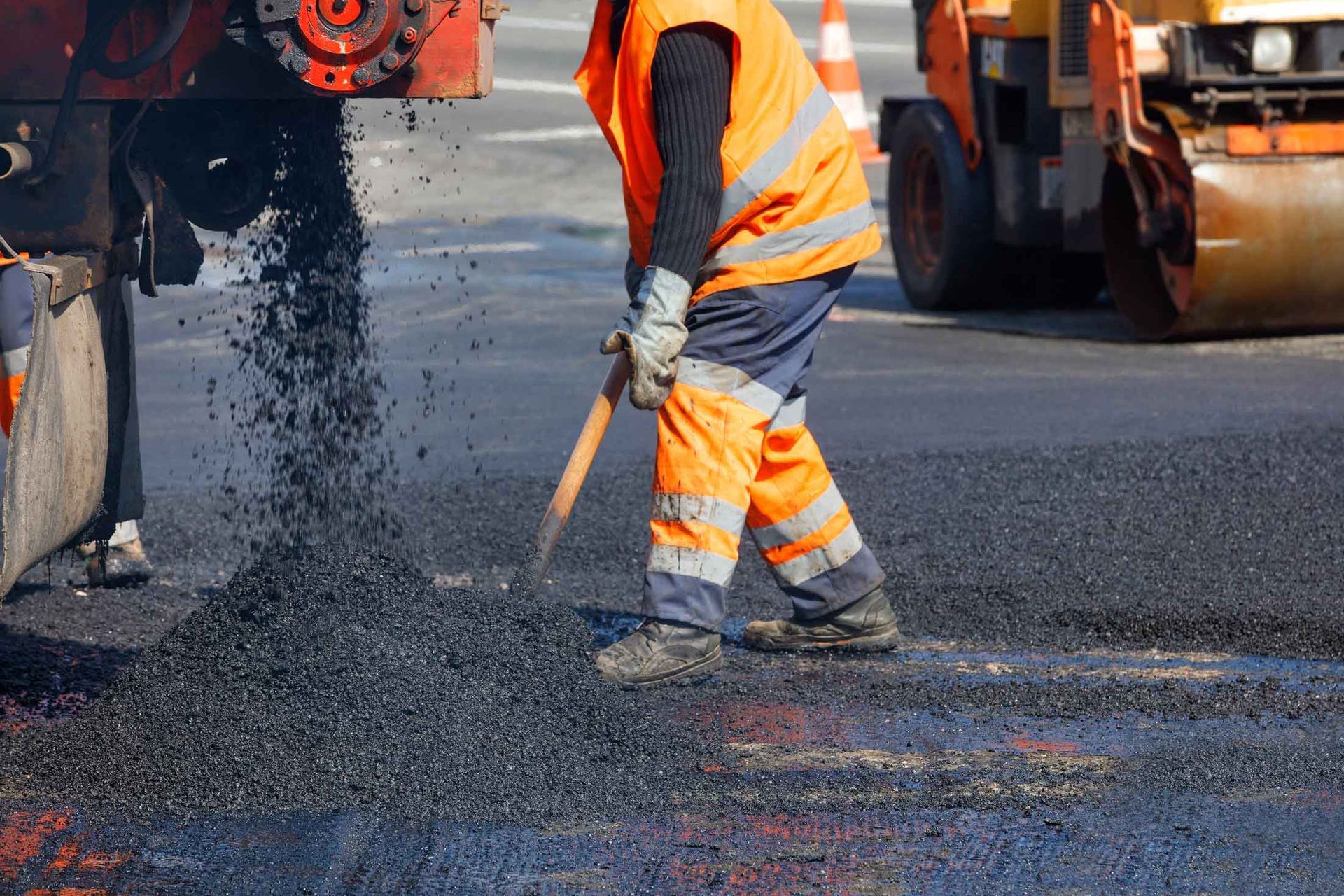 Worker spreads hot asphalt while paving machine pours material during road repair.