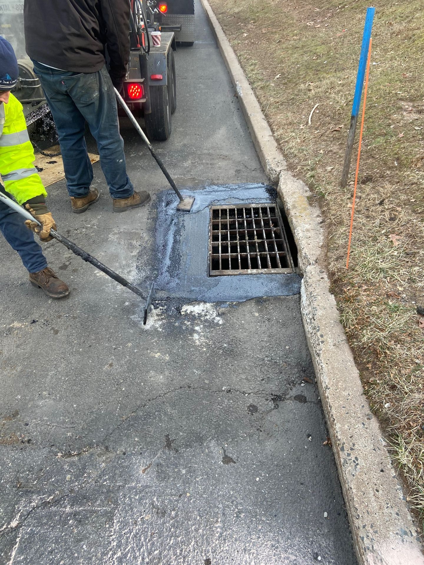 Workers repairing asphalt around a storm drain on a concrete curb, with a vehicle in the background.