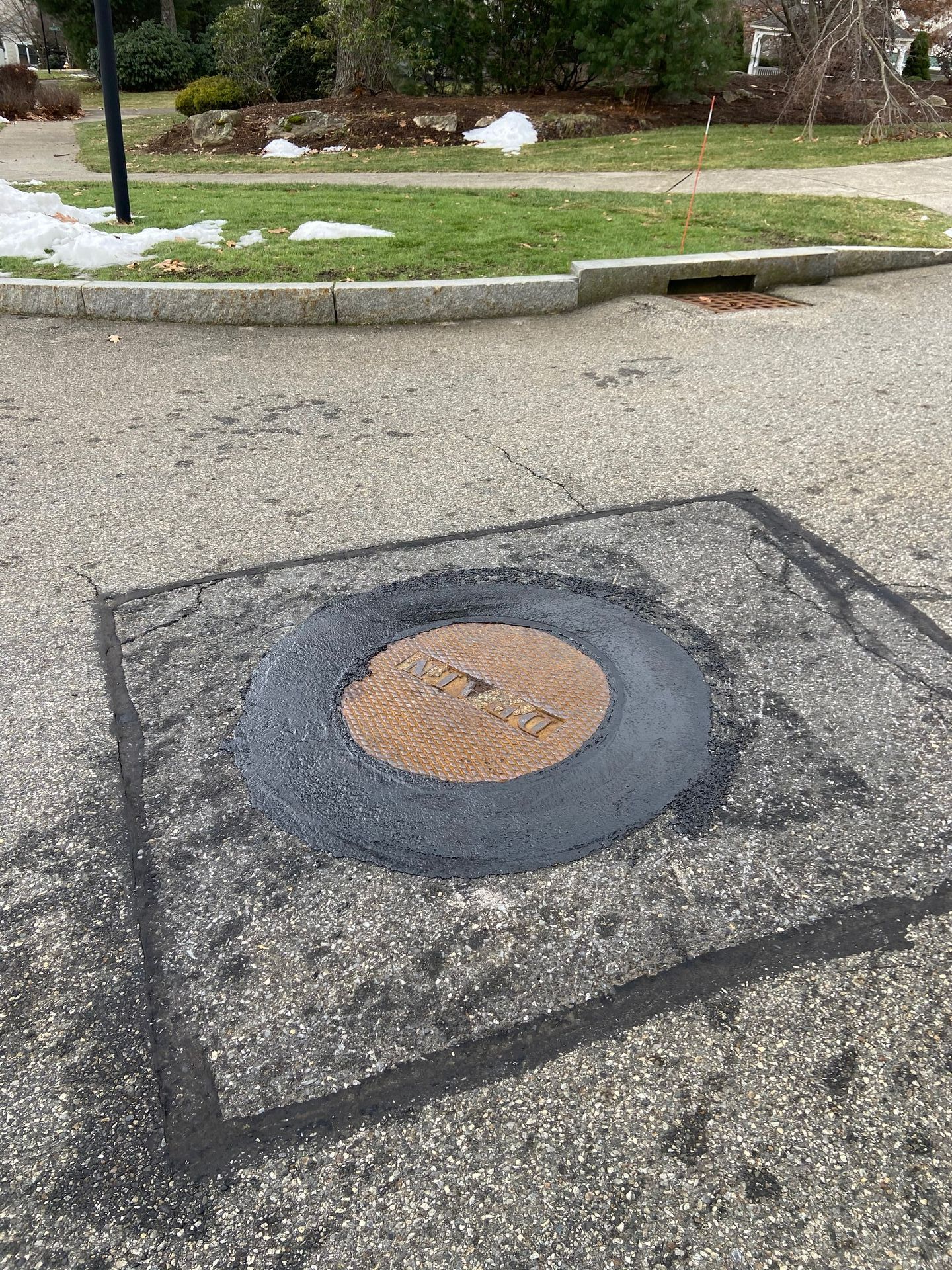 Asphalt road patch around a manhole cover. The patch is rectangular and painted black.
