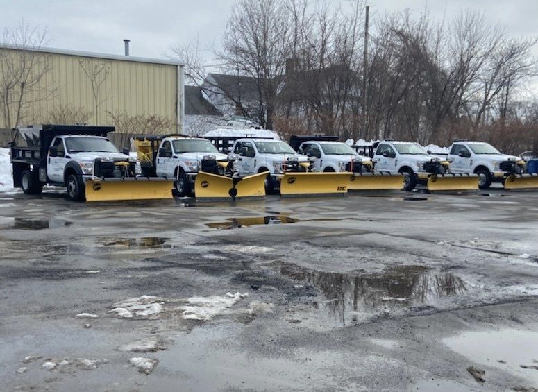 Six white pickup trucks with snow plows parked on a wet, icy lot.