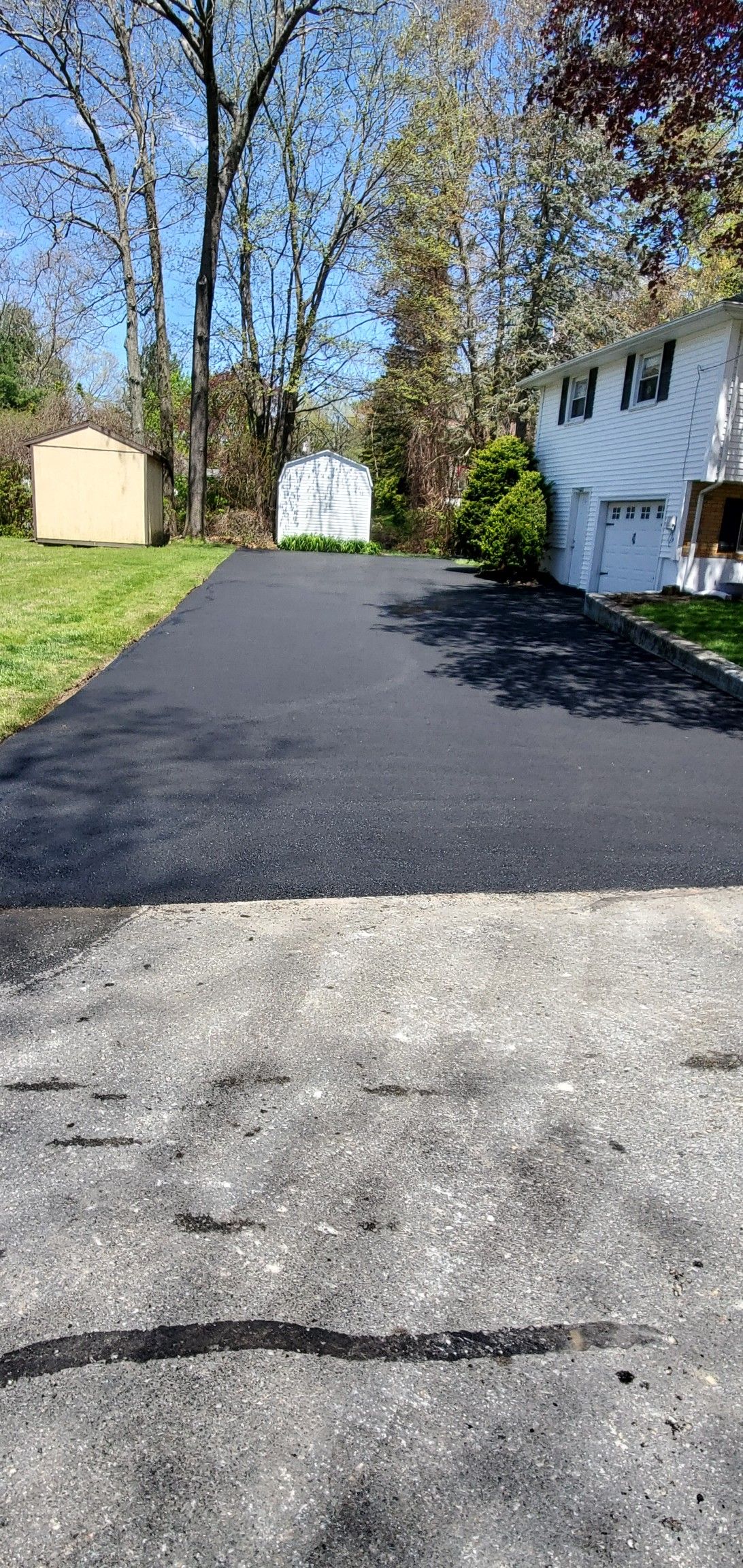 Driveway with dark asphalt leading to a white house and a small yellow shed, sunny day.