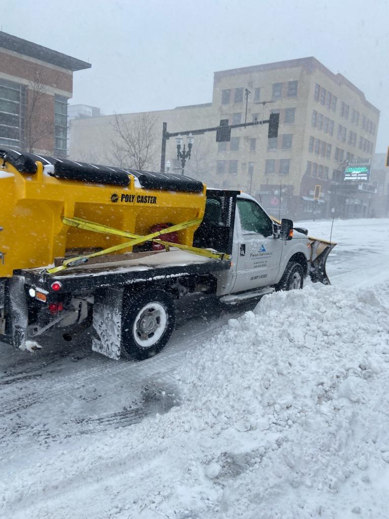 Snowplow clearing snow on a street in front of buildings during a snowstorm.