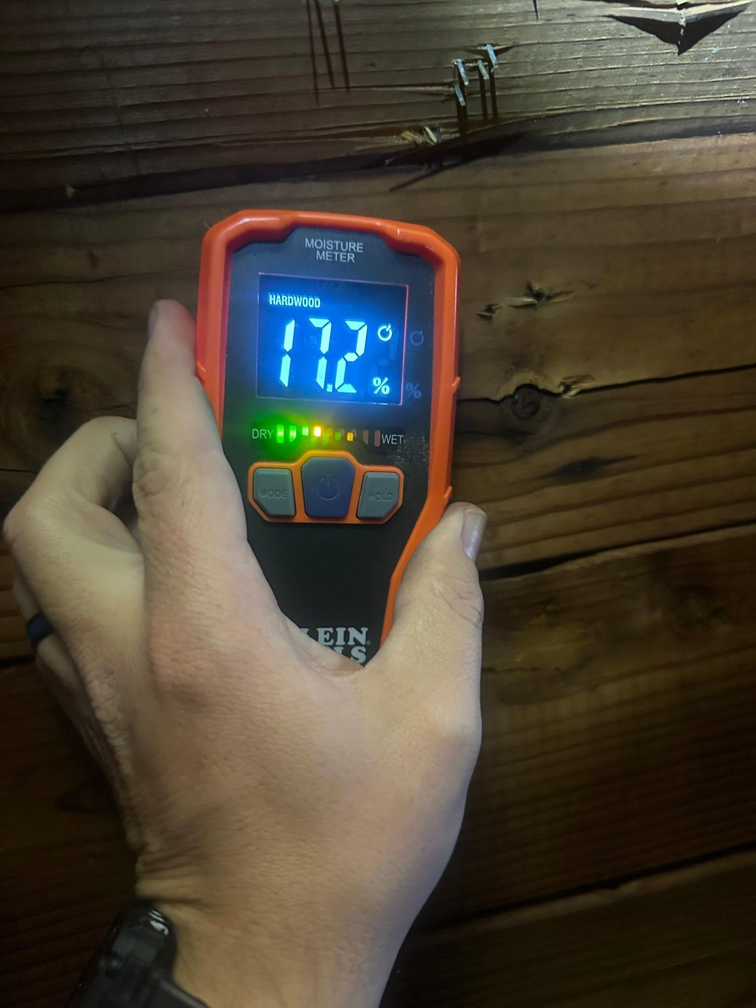 A person uses an orange moisture meter on a wooden surface, displaying 17.2°C and 7%.