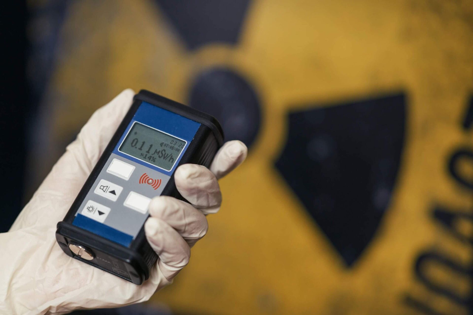 Hand holding a radiation detector in front of a yellow radiation hazard symbol.
