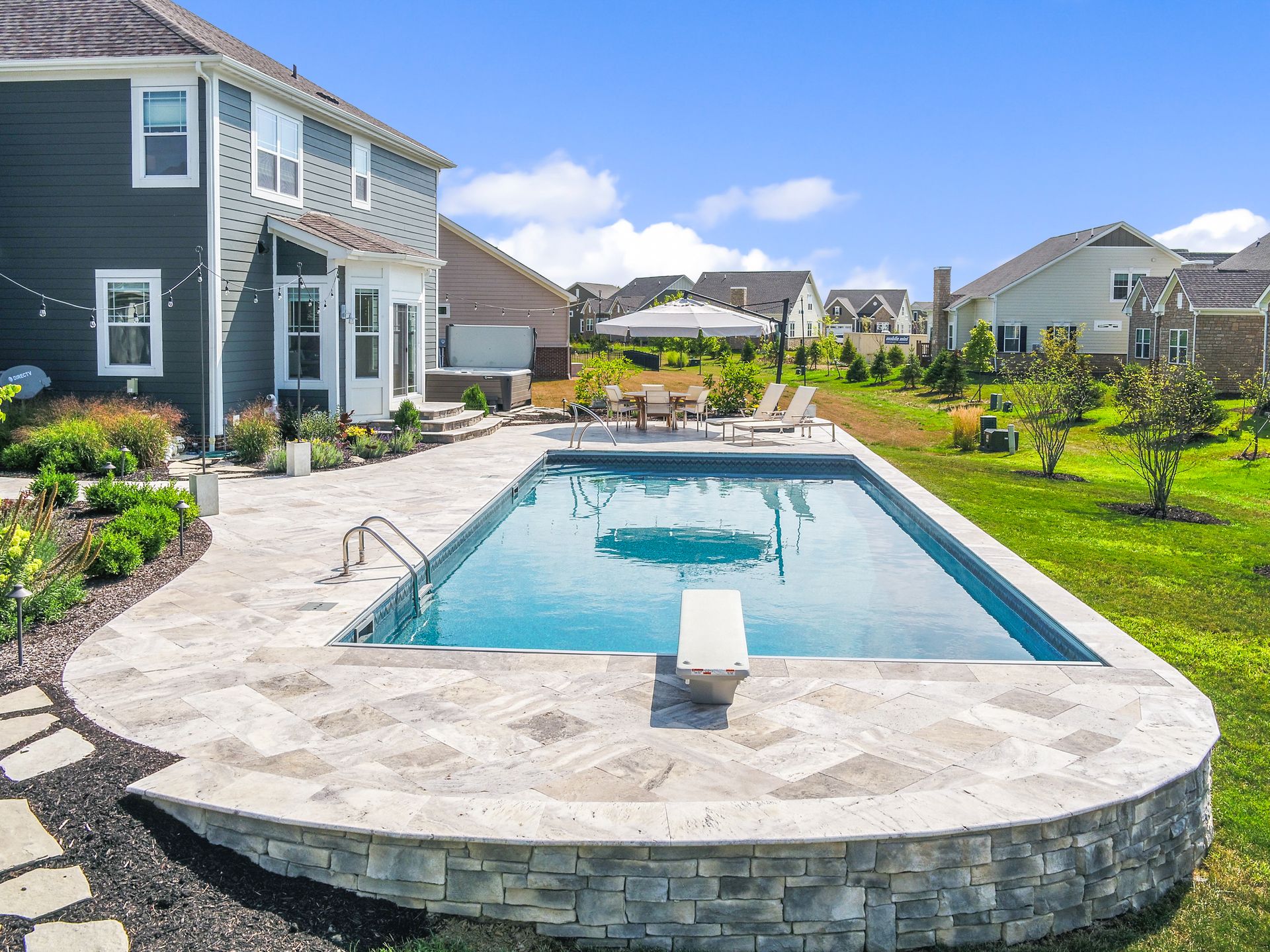 Backyard pool with diving board surrounded by stone patio, house in the background, blue sky.