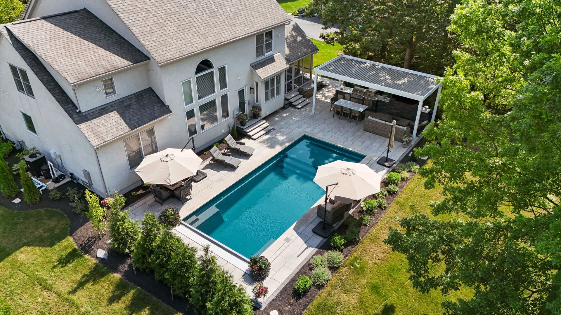 Backyard pool with diving board surrounded by stone patio, house in the background, blue sky.