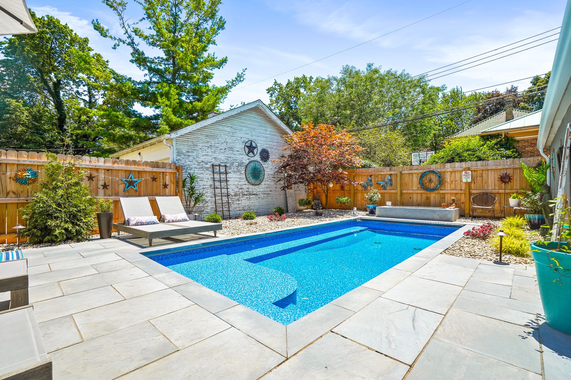A backyard pool with a patio, lounge chairs, and a wooden fence. Sunny day with lush greenery and a small building.