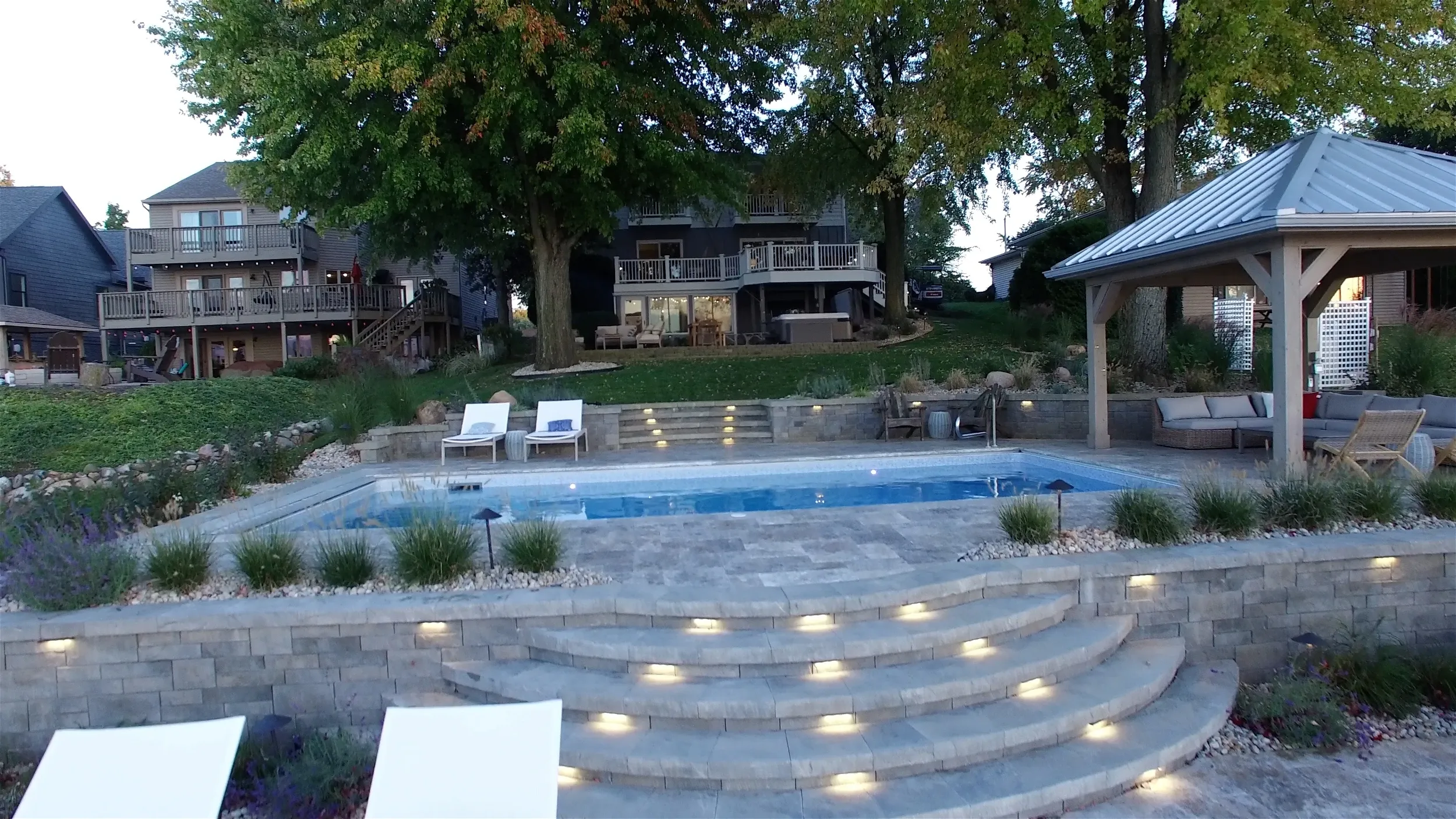 Stone steps lead to a pool with a gazebo and houses in the background. The pool is surrounded by greenery.