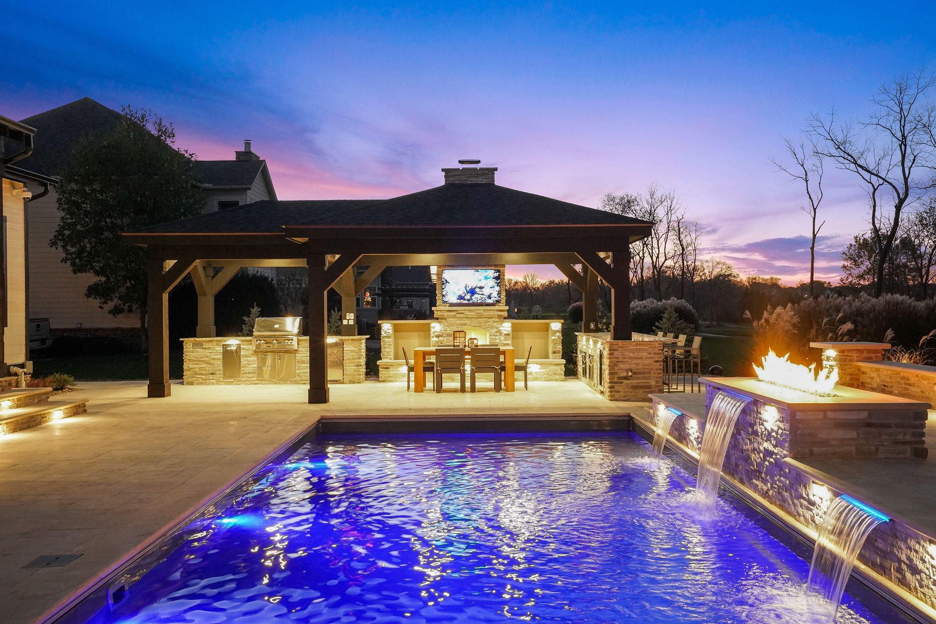 A luxurious backyard with a pool, outdoor kitchen, and a lit fire pit, all set against a dusk sky.