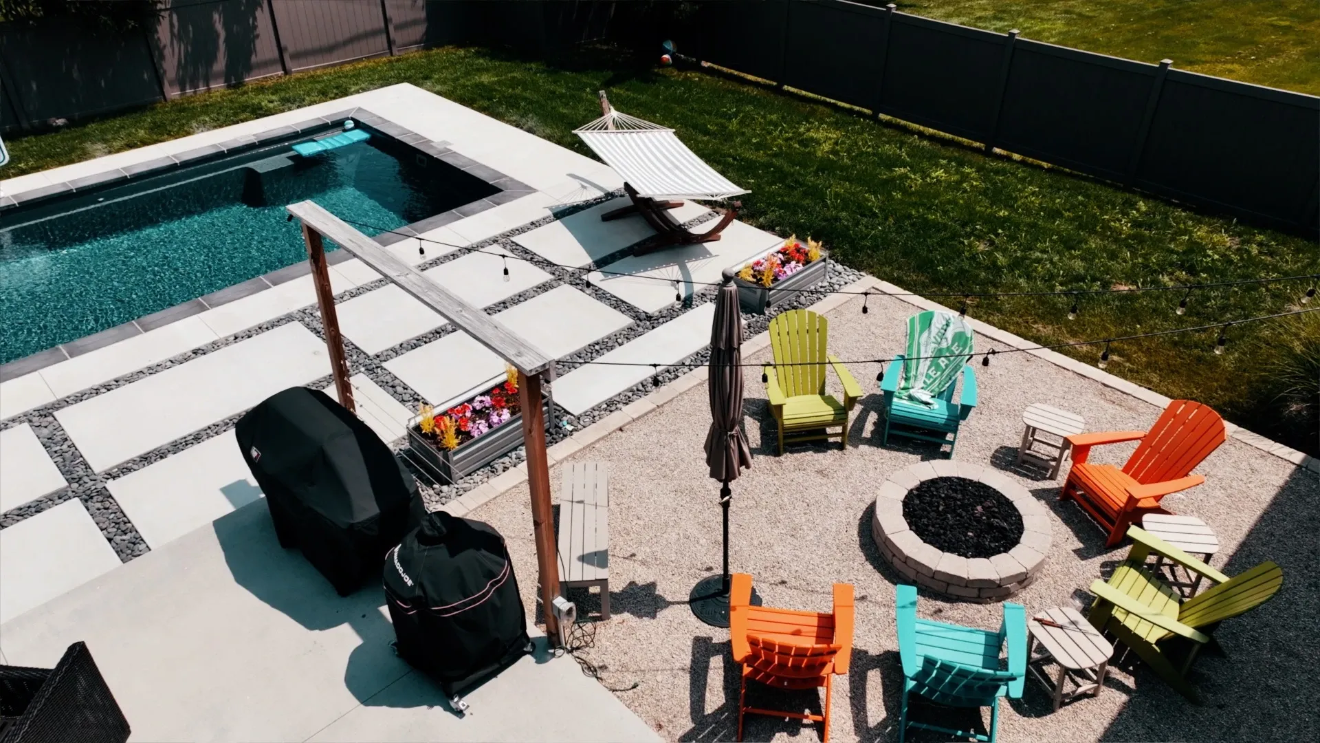 An aerial view of a backyard with a pool, fire pit, and colorful Adirondack chairs arranged in a circle.