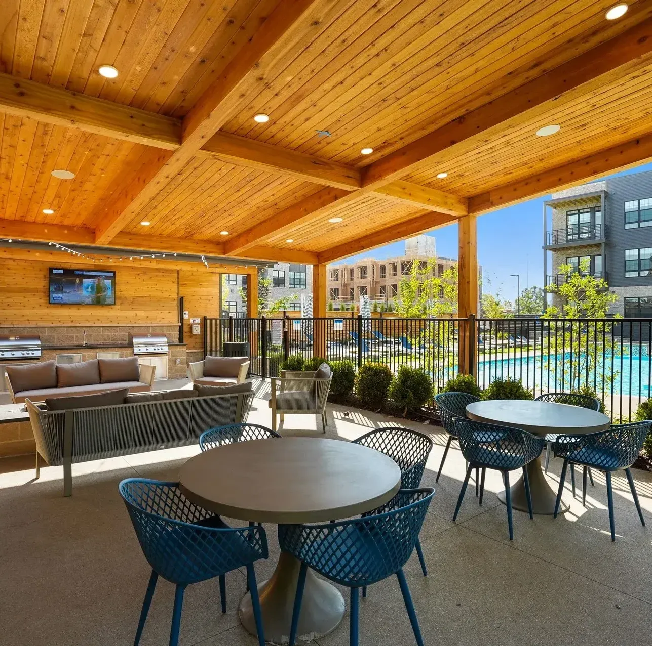 Outdoor lounge area with seating, tables, and a covered wooden ceiling, overlooking a pool and adjacent building.