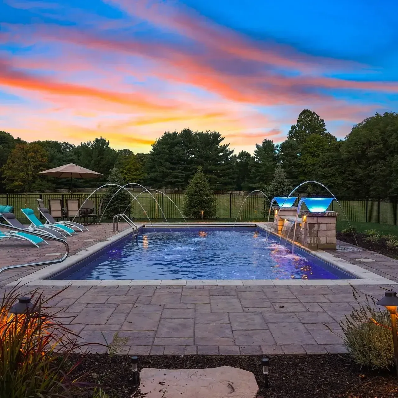 Backyard pool with fountains and blue lights, surrounded by a stone patio and lounge chairs, at sunset.