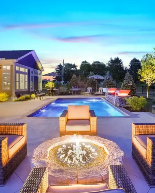 A backyard with a pool, seating around a fire pit, and a house in the background at dusk.