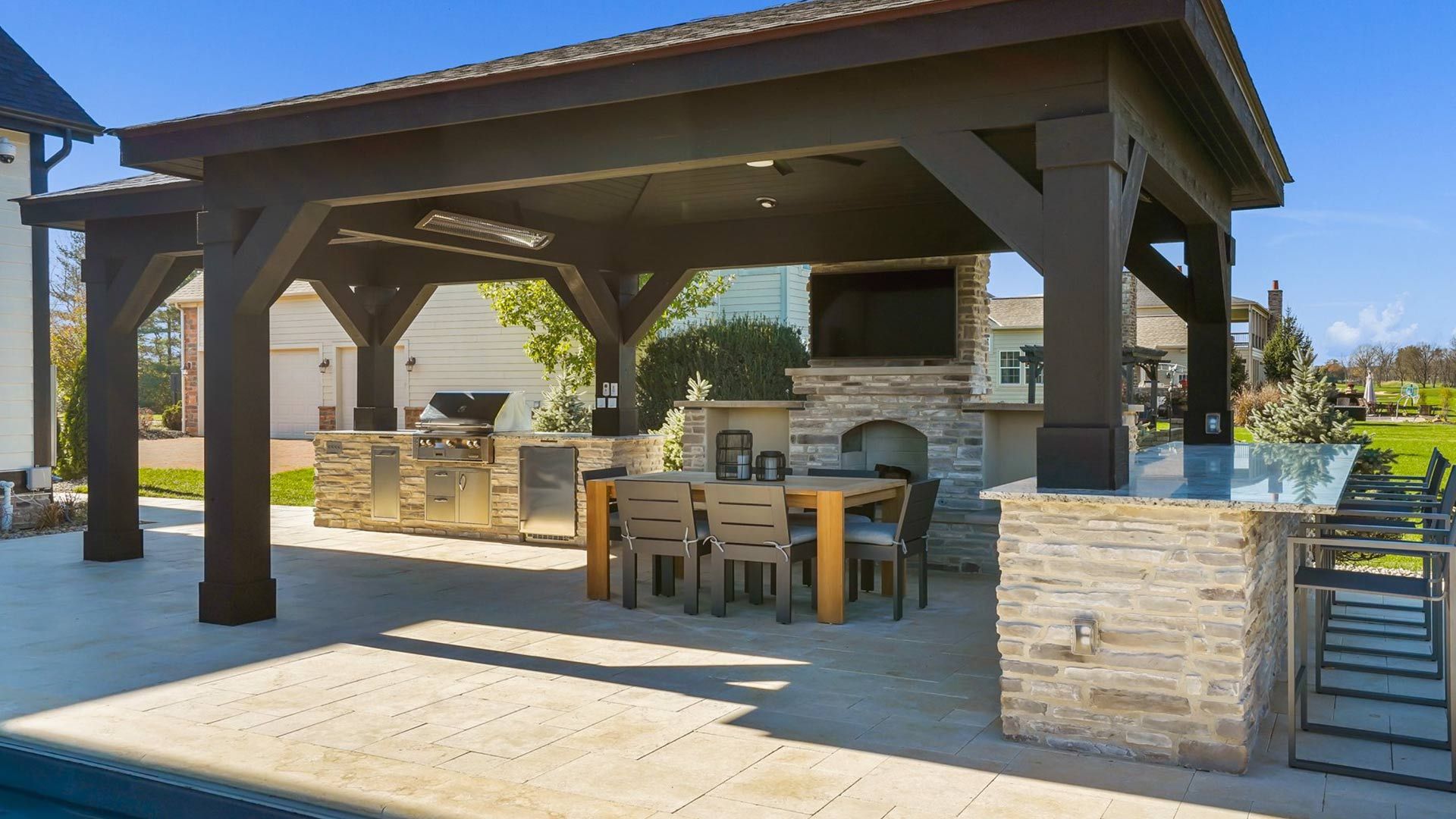 Outdoor kitchen and dining area under a dark wooden pergola with stone accents. Features a grill, fireplace, table, and bar.