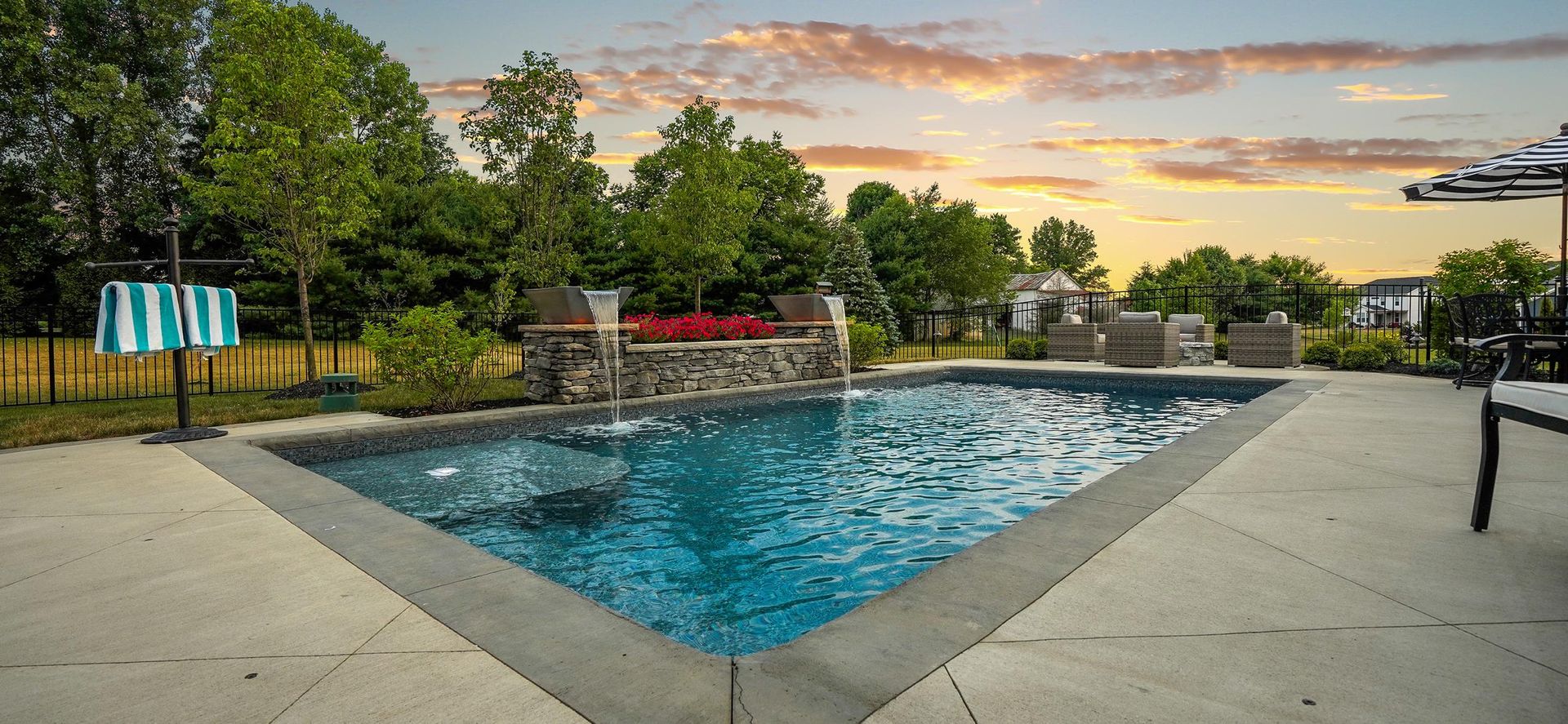 A swimming pool with a stone wall, surrounded by a concrete patio, and trees in the background. A towel rack with striped towels is in the foreground.