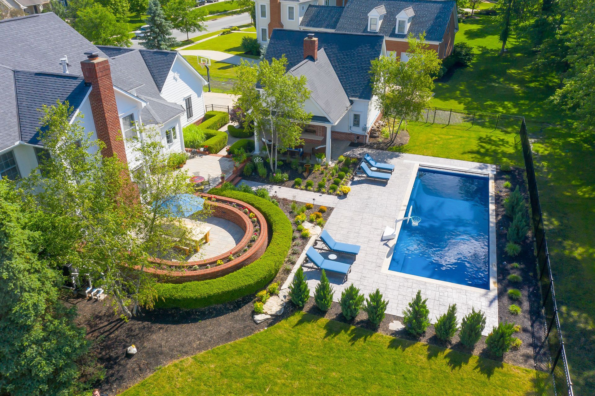 Aerial view of a luxurious backyard with a pool, patio, and manicured landscaping; a large house is in the background.
