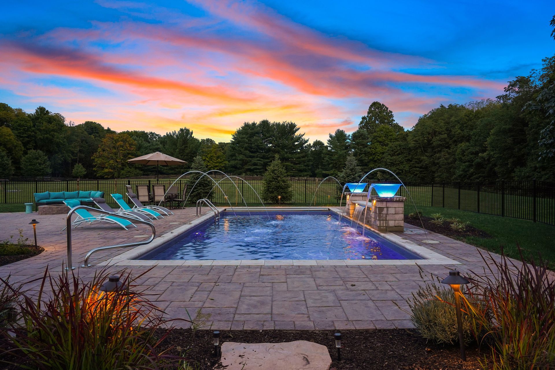 Backyard pool at sunset with lounge chairs, stone patio, and colorful sky.