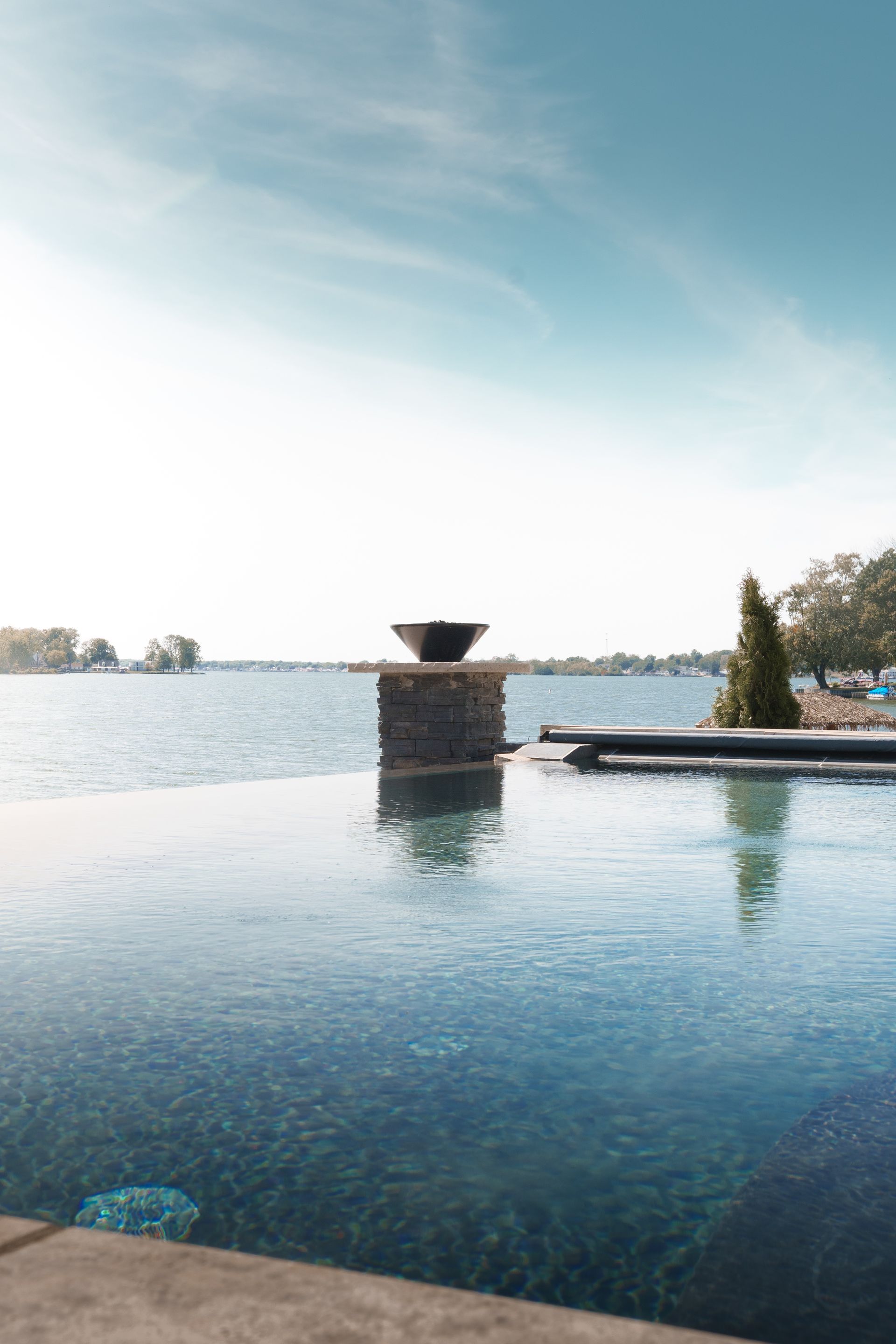 An infinity pool overlooks a lake on a sunny day. A stone pillar with a fire bowl sits in the pool's center.