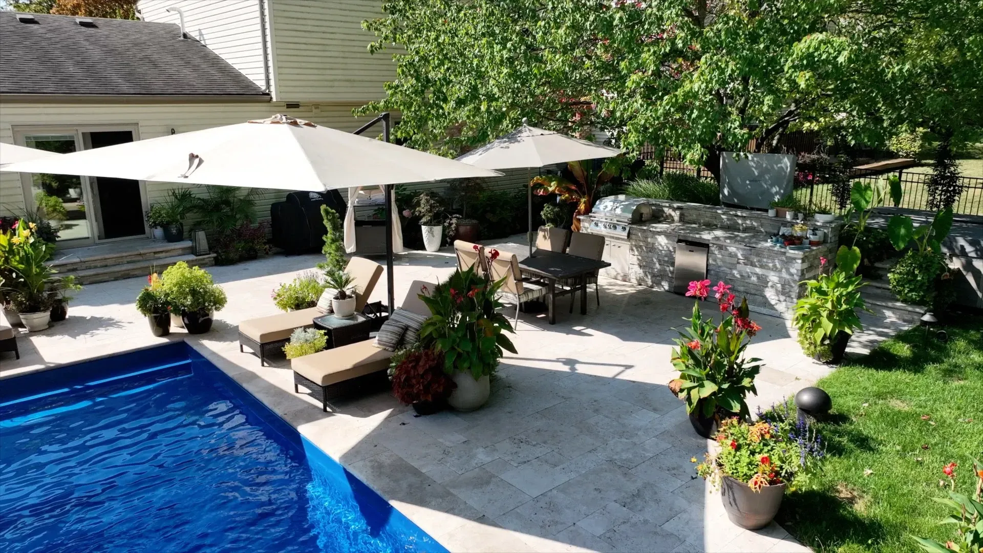 Backyard patio with pool, lounge chairs under umbrellas, outdoor kitchen, and lush greenery.