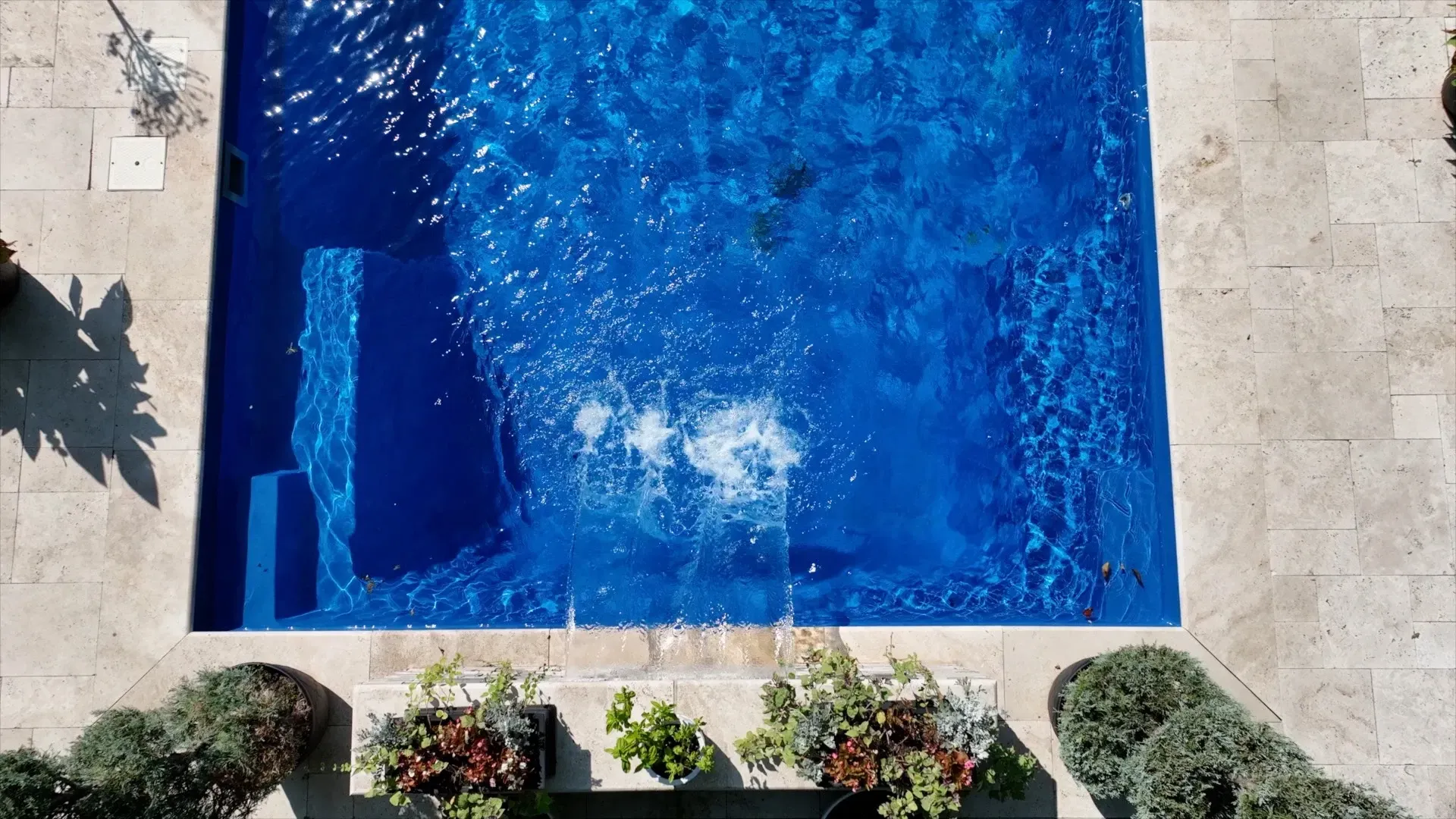Overhead view of a blue rectangular pool with steps, bubbles indicating someone has just entered the water, surrounded by light-colored stone and greenery.