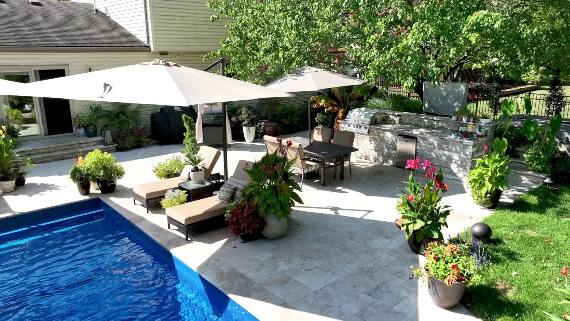 Backyard patio with pool, lounge chairs, dining table, and built-in outdoor kitchen. Lush greenery surrounds the paved area under umbrellas on a sunny day.