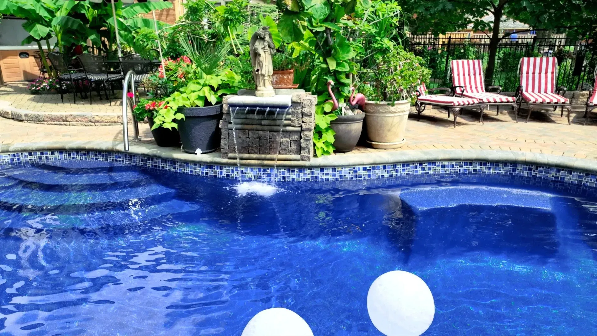 A sparkling blue pool with steps, a waterfall, and lounge chairs.  Greenery surrounds the pool in a sunny outdoor setting.
