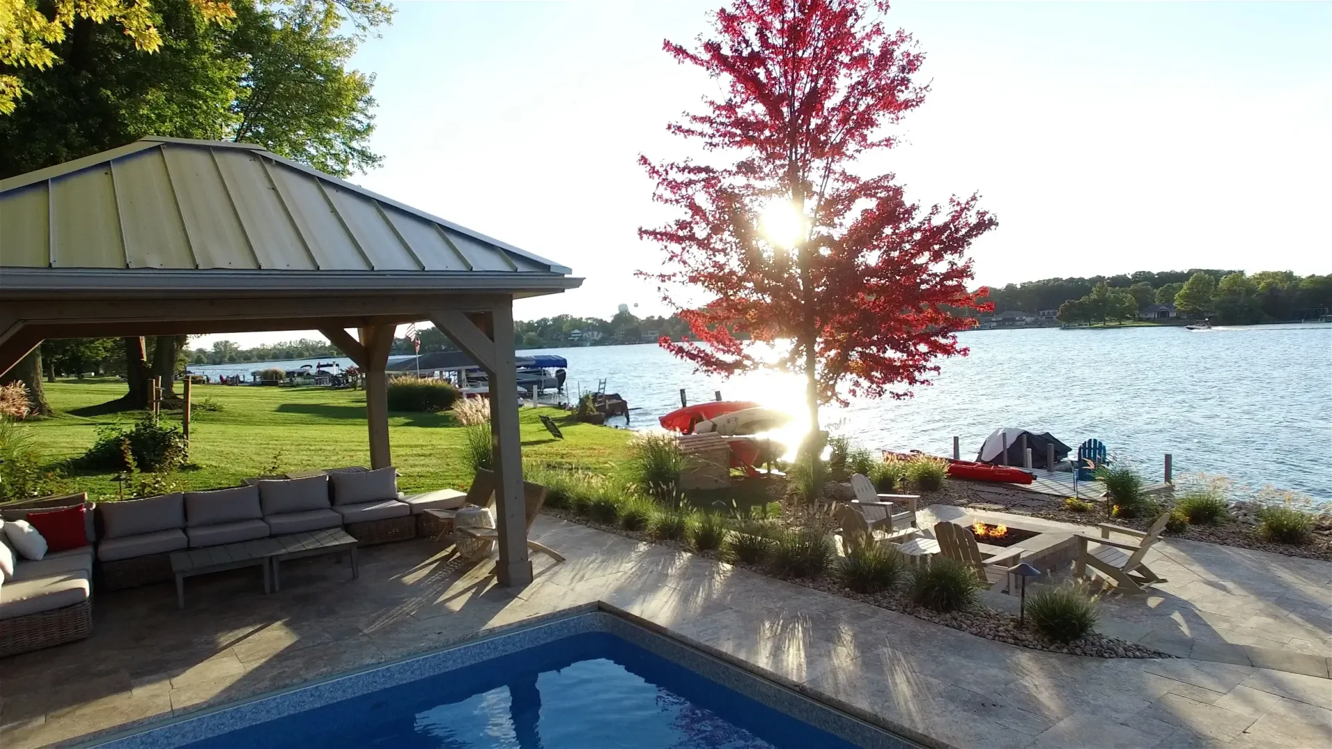 A lakeside scene with a gazebo, pool, and fire pit. Sunlight shines through a vibrant red tree, reflecting off the water.