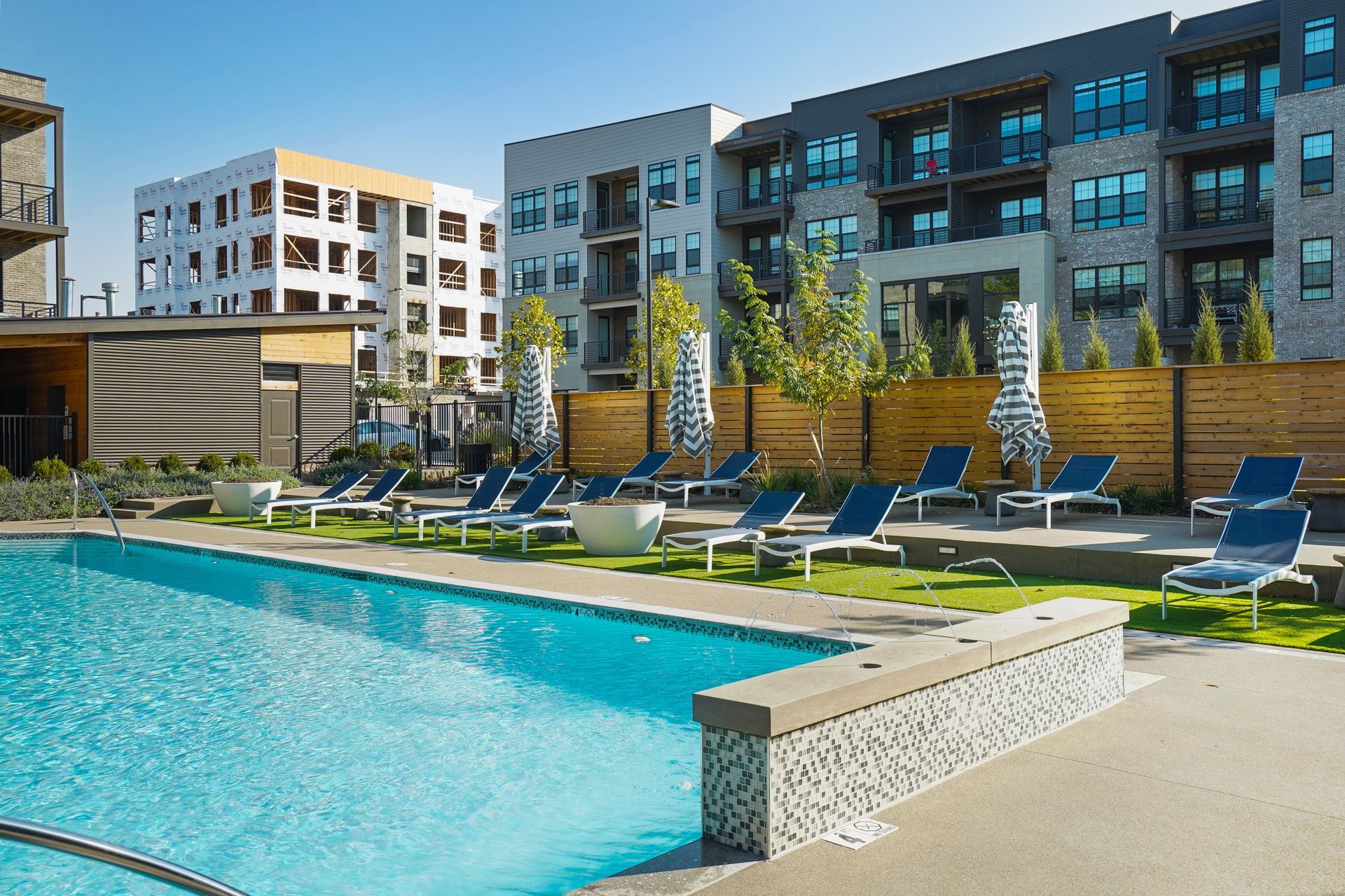 Pool area with lounge chairs, and apartment buildings in the background. Blue pool water, tan patio, and a wooden fence.
