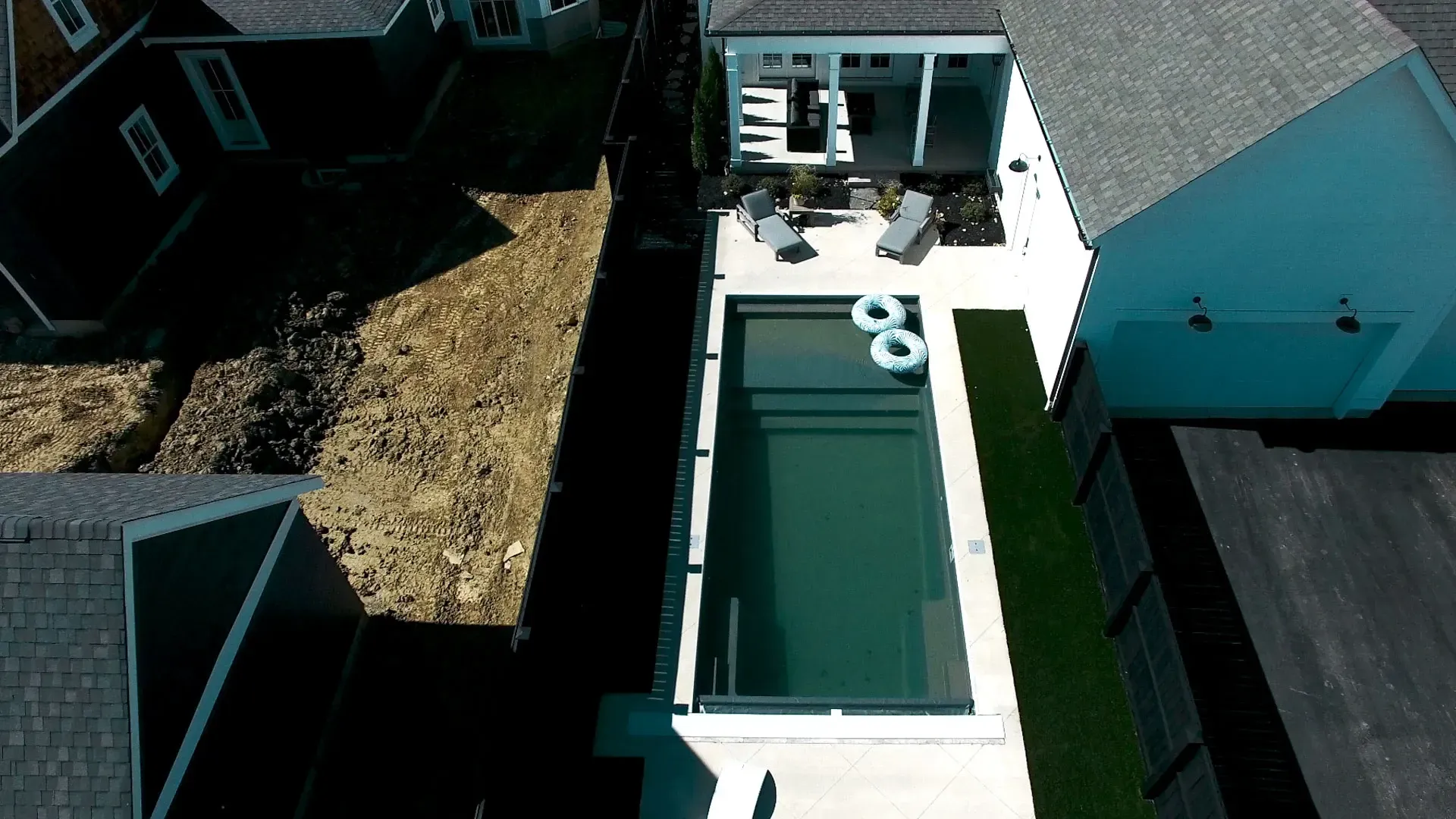 Aerial view of a rectangular swimming pool between two houses, with floaties in the water and patio furniture on the deck.